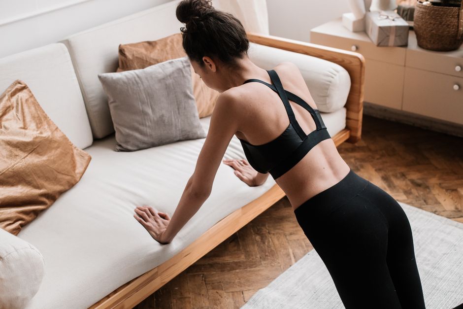 An active adult woman performs push-ups indoors using a sofa for support, emphasizing a healthy lifestyle.