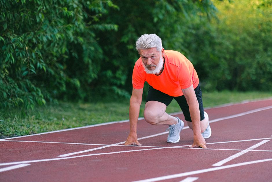 Full body of confident focused senior male ready for sprint on racetrack in stadium near green bushy plants