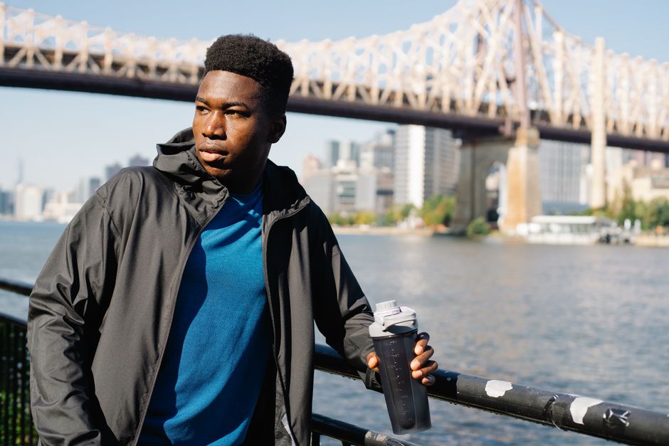 Man in black jacket holding a water bottle on a bridge with city skyline in background.