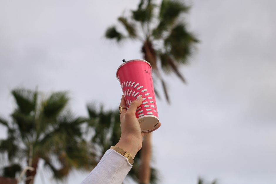 A person holds a vibrant pink cup outdoors with palm trees in the background.