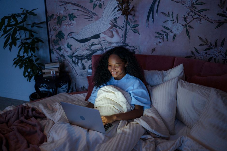 Teenage girl smiling while using a laptop in bed under soft lighting at night, enjoying her time online.