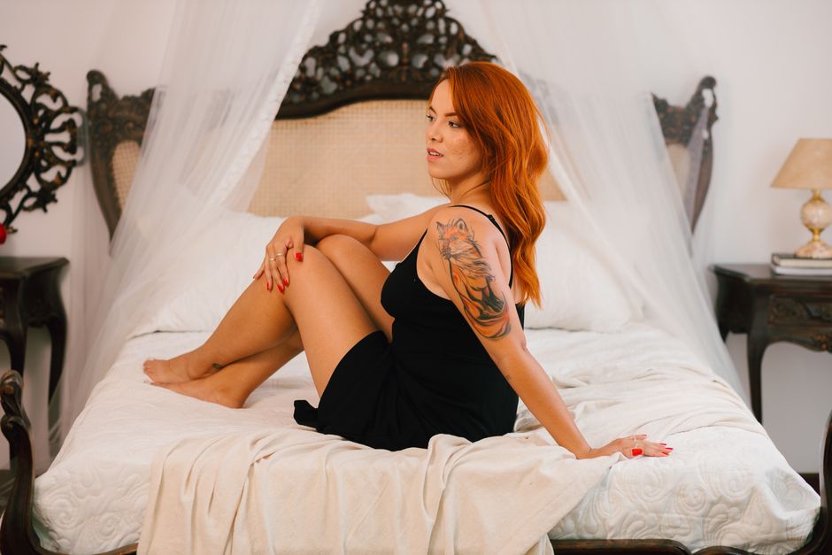 A red-haired woman with a tattoo sits on a luxurious bed, embracing a calm ambiance.
