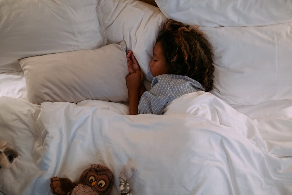 A young girl peacefully sleeping under soft, white bedding with a plush owl nearby.
