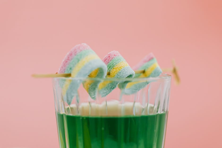Colorful candy skewers dipped in green beverage against pink backdrop.